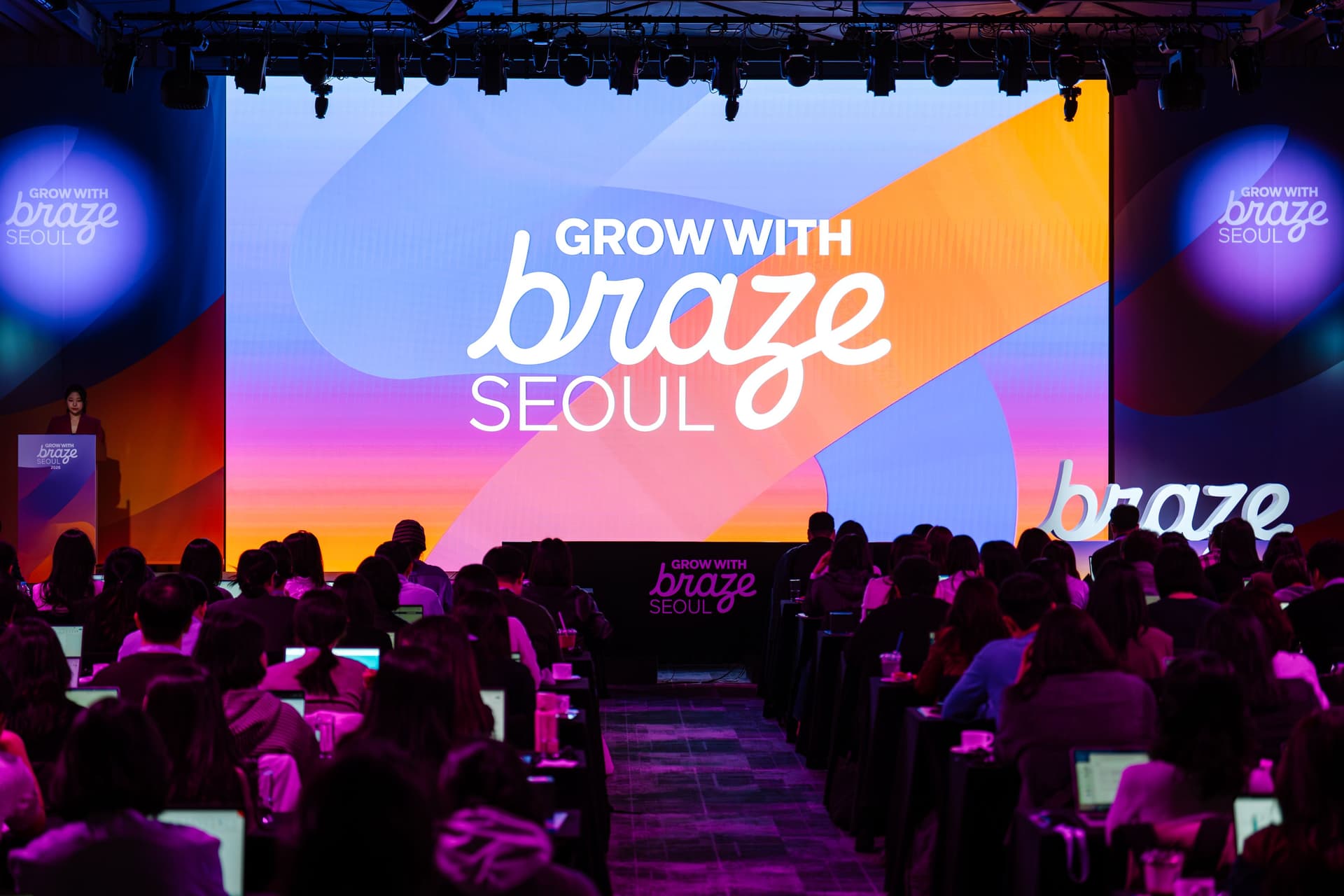 A conference hall filled with attendees facing a large screen displaying "GROW WITH braze SEOUL" and a speaker at a podium.