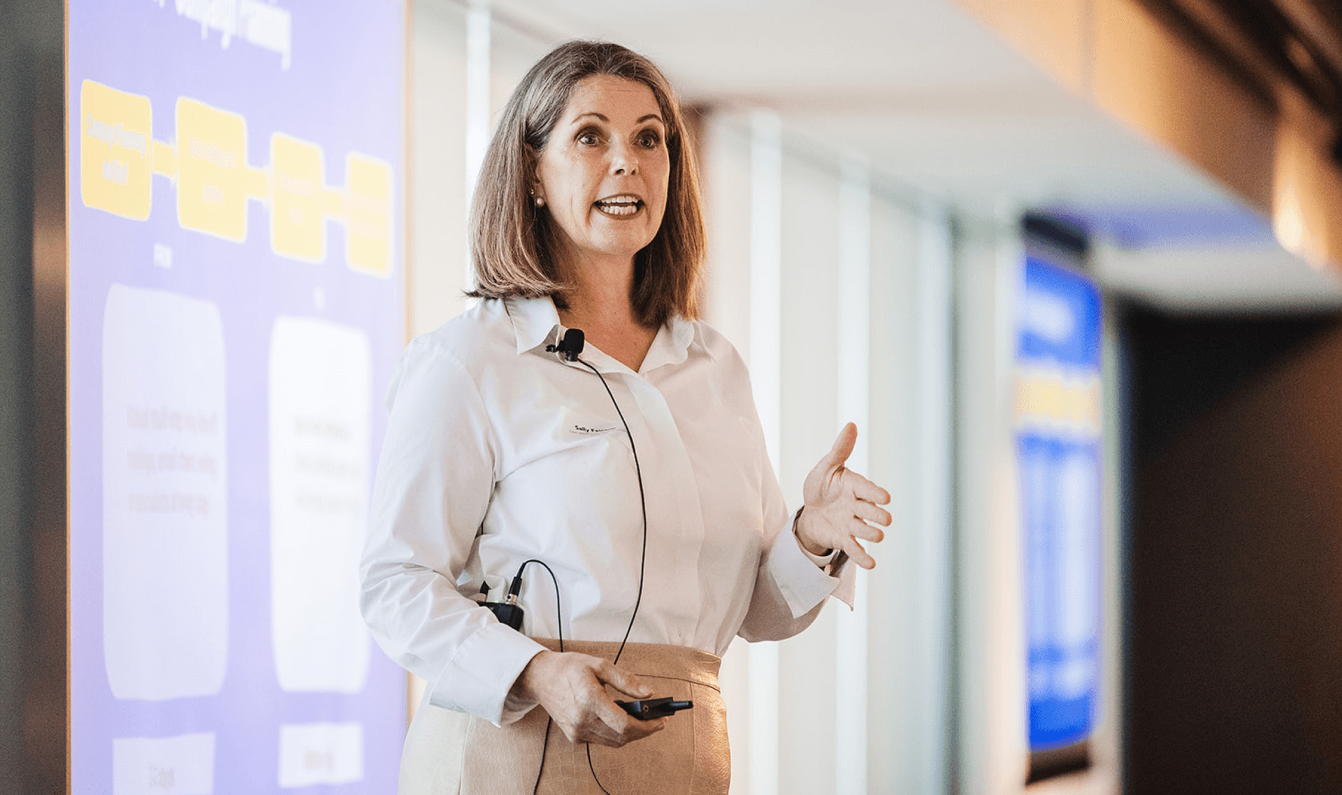 A woman in a white shirt and tan skirt presents with a microphone, gesturing and holding a clicker.