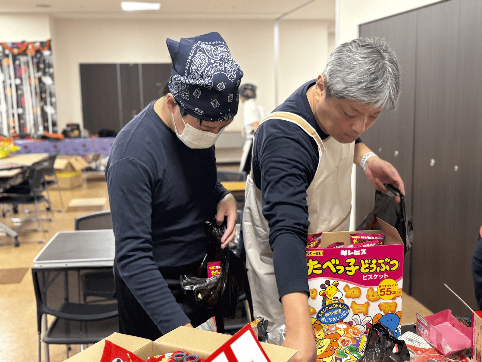 Two men pack snack boxes and other items into bags on a table.