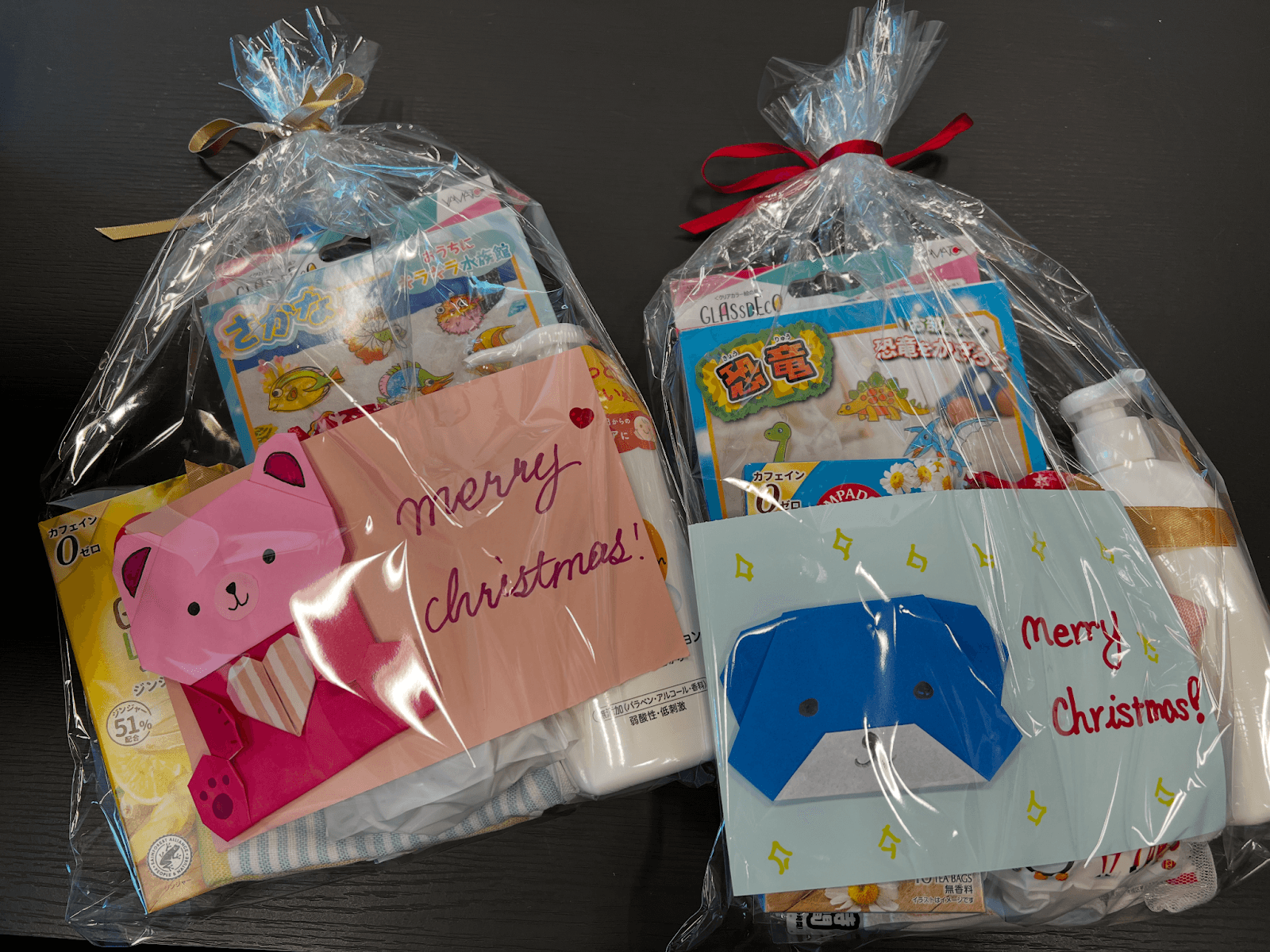 透明なクリスマスギフトバッグ2つに、金と赤のリボンが結ばれ、折り紙の動物のカードと、お風呂用おもちゃや工作キットなどの様々なプレゼントが入っている。
