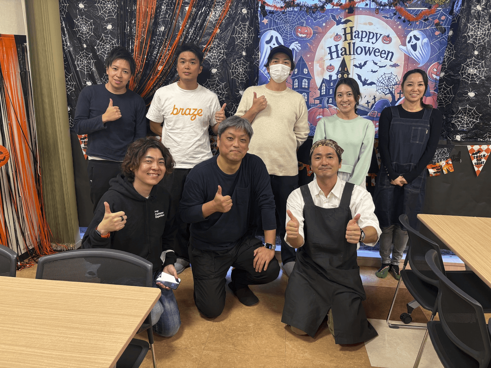 Eight people, some wearing aprons, smile and give thumbs-up in a Halloween-decorated room.
