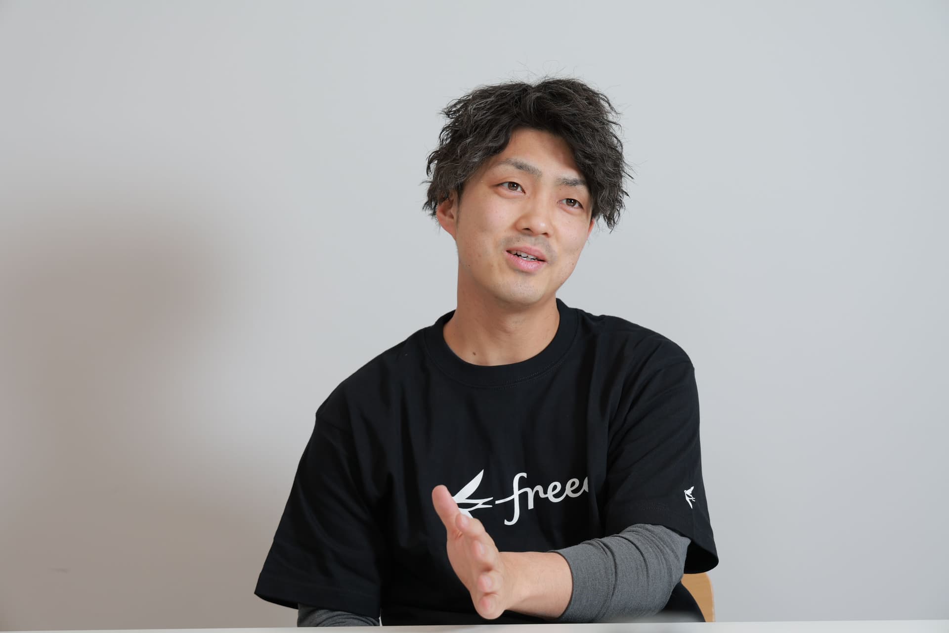 黒く縮れた髪をした男性が、「free」のロゴが入った黒いTシャツを着て、話しながら身振りを交えている。