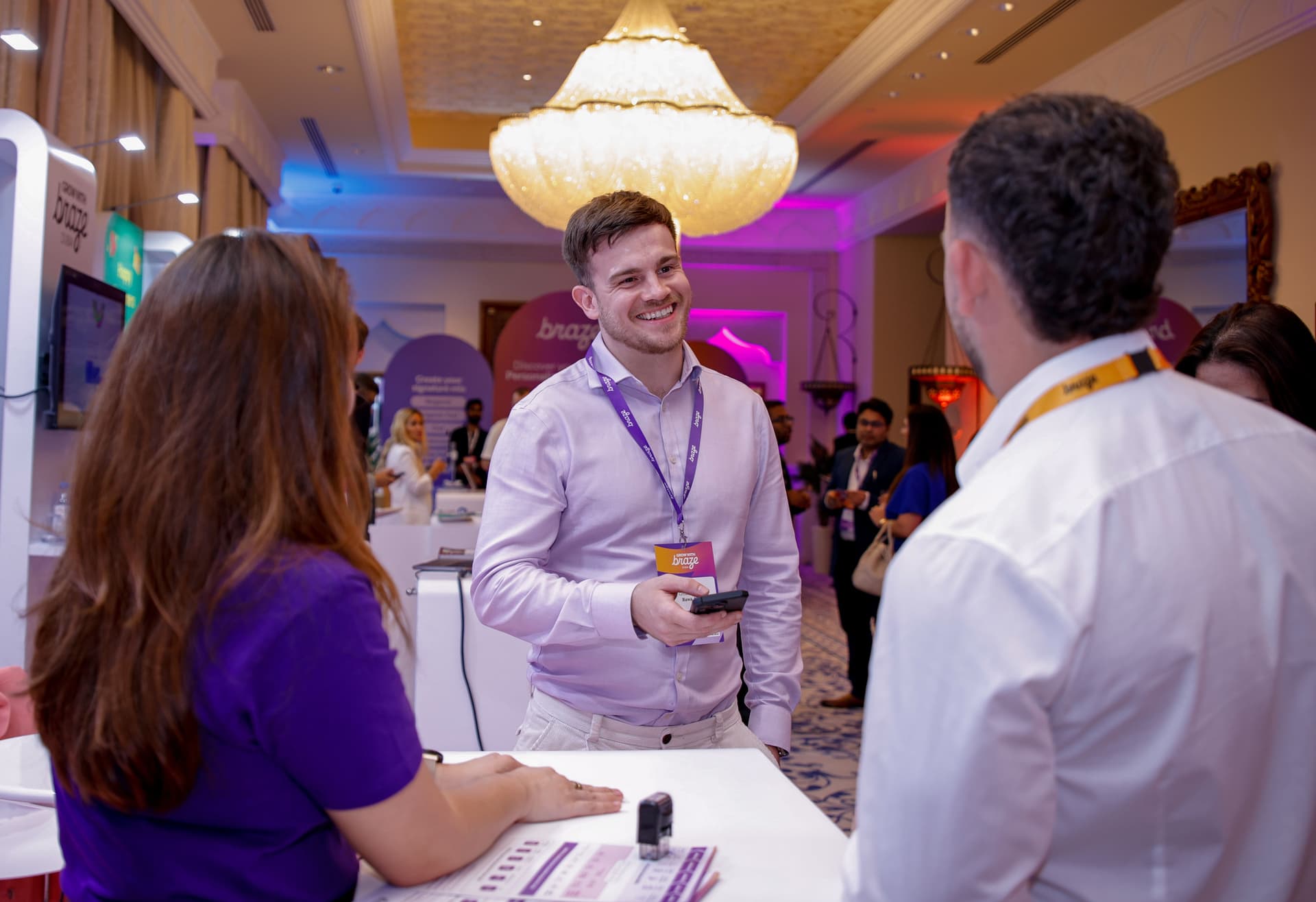 A smiling man in a purple shirt talks to another attendee at a bustling indoor event.