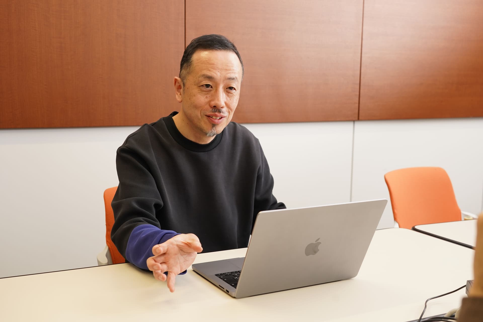 暗い色のスウェットシャツを着た男性がテーブルに腰かけ、ノートパソコンを前にして話しながら身振り手振りで説明している。