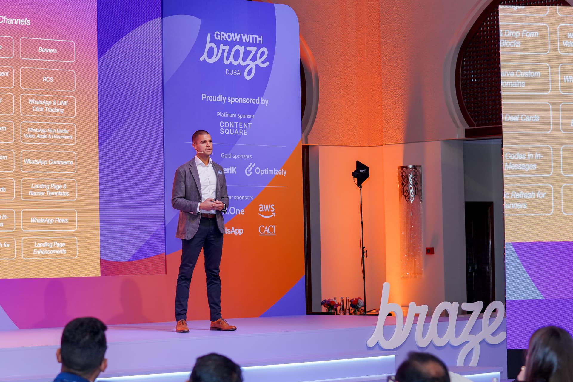 A man presents on a stage at a "Grow with Braze Dubai" event, with feature lists and sponsor logos on screens behind him.