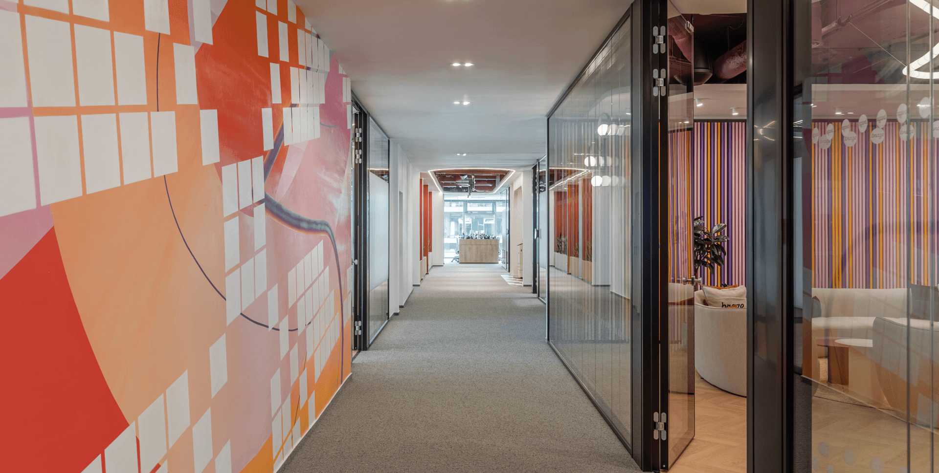 A modern office hallway with a colorful abstract mural on the left and glass-fronted rooms on the right.