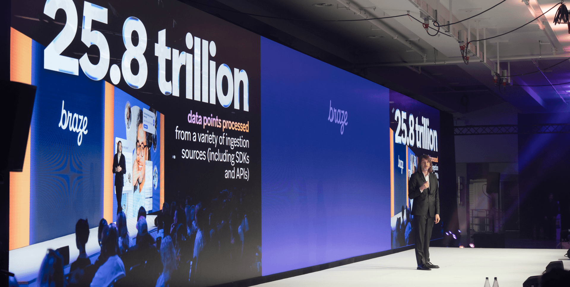 A speaker presents on a stage with large screens displaying "25.8 trillion" and "Braze" to an audience.