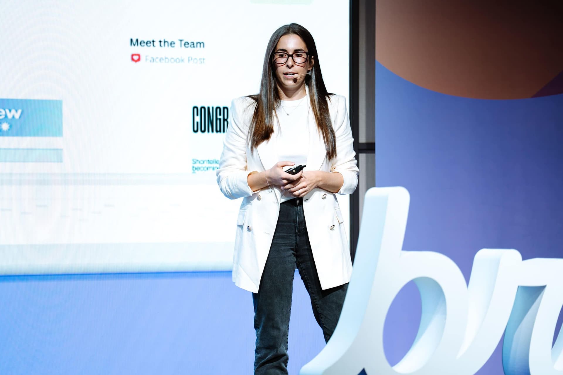 A woman in a white blazer presents on stage, holding a remote, with a screen and large white decorative letters.