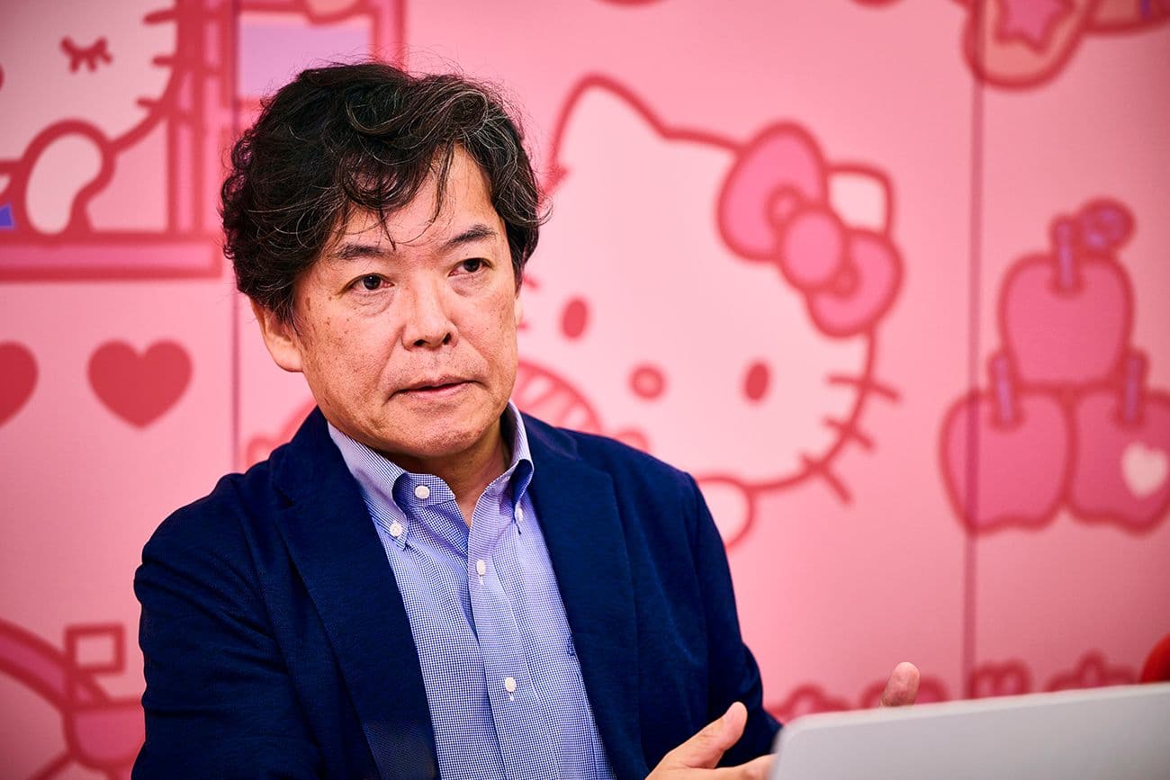 青いシャツとブレザーを着た男性(サンリオ デジタル事業本部 デジタル事業開発部 CX推進課 顧客体験プラットフォーム戦略推進担当 田口 歩氏)が、ハローキティのシルエットで飾られたピンクの壁の前に座っている。