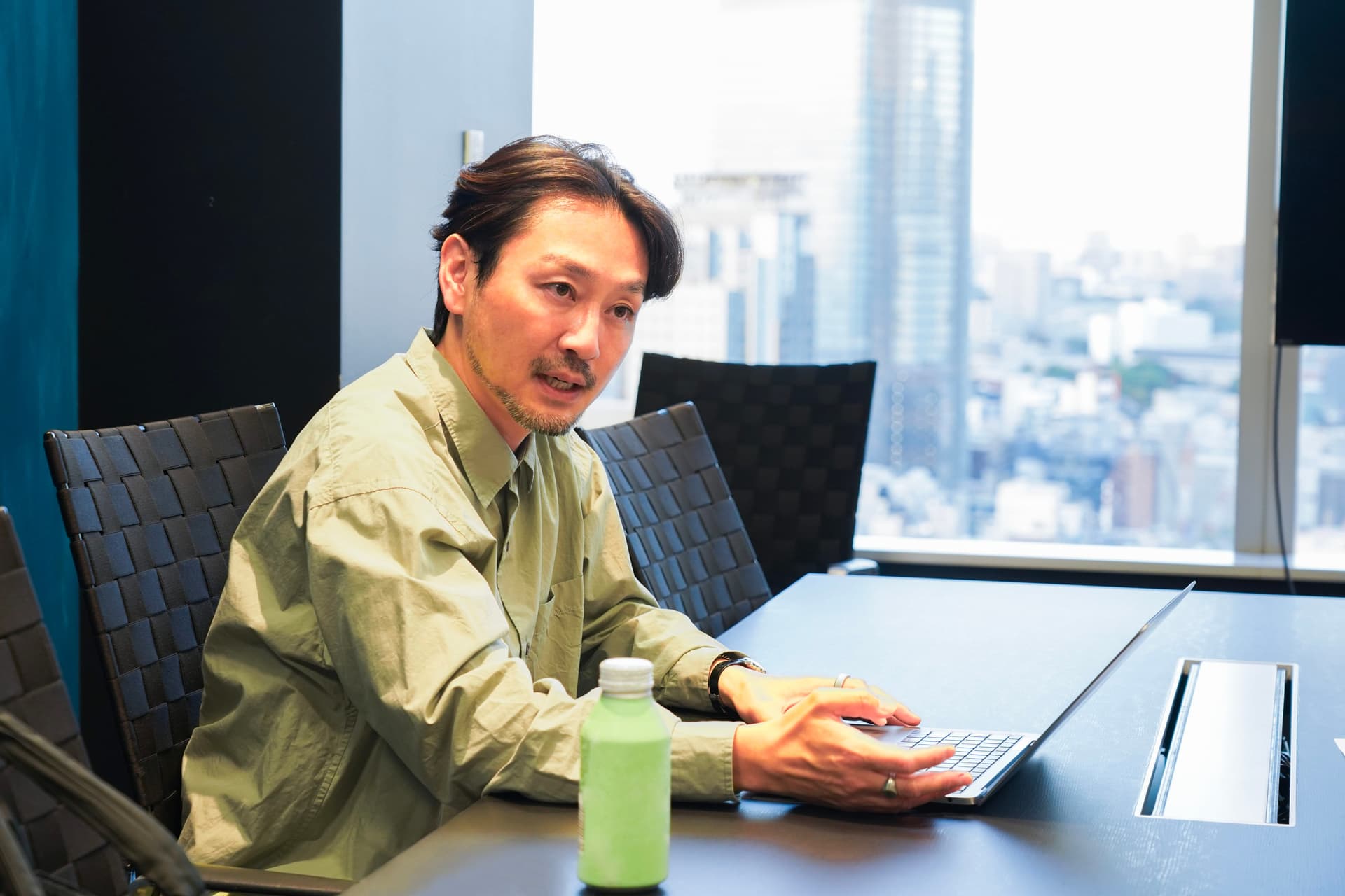 A man sits at a conference table using a laptop, with a cityscape visible through a window.