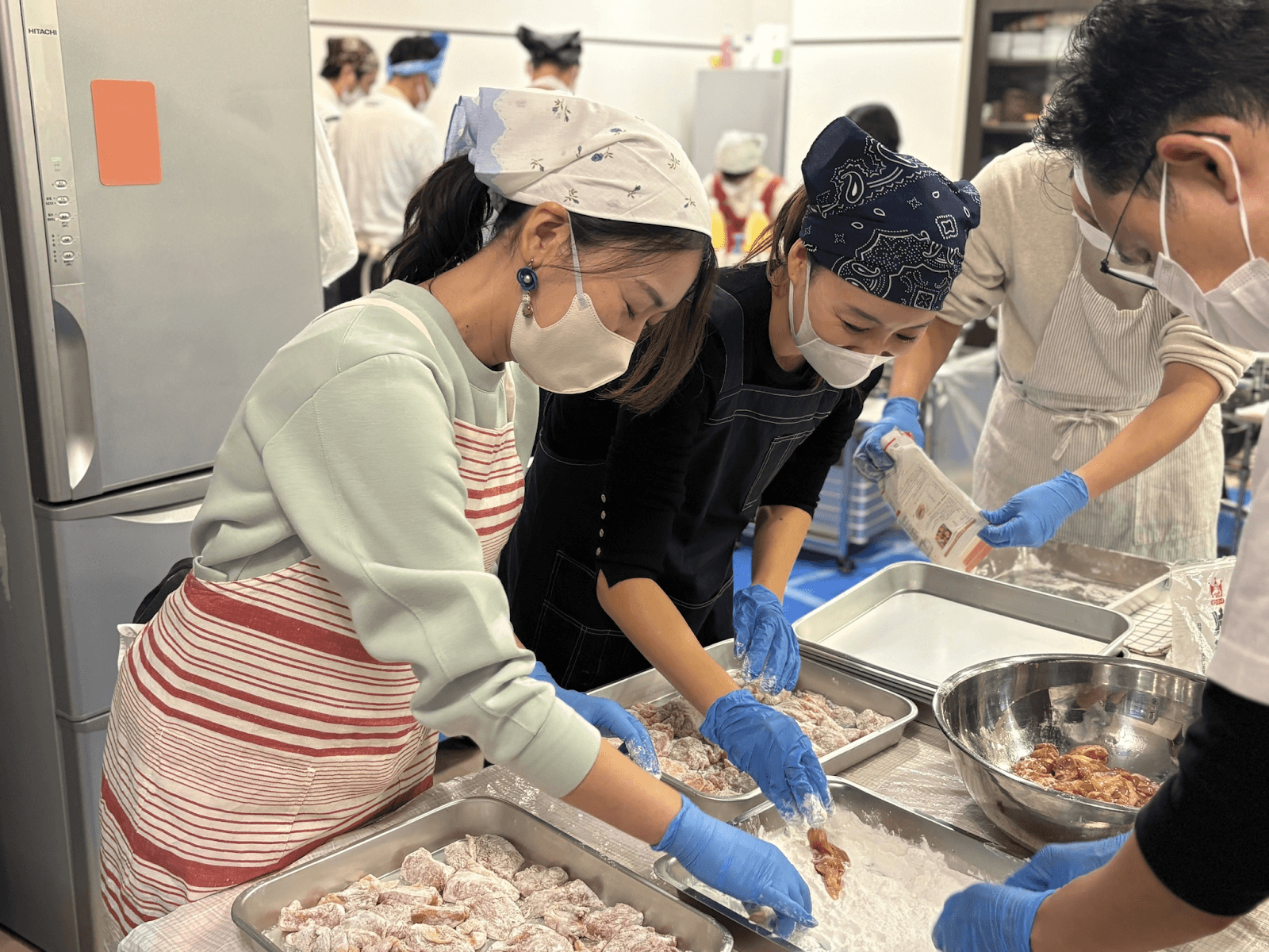 People in masks and gloves breading chicken in a kitchen setting.
