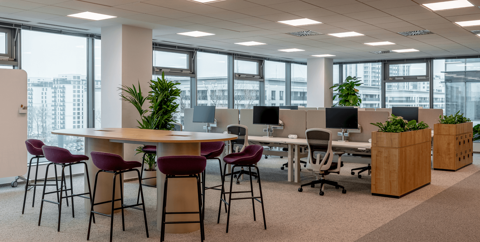 A modern open-plan office featuring a collaborative high-top table with stools, individual workstations, and large windows with a city view.