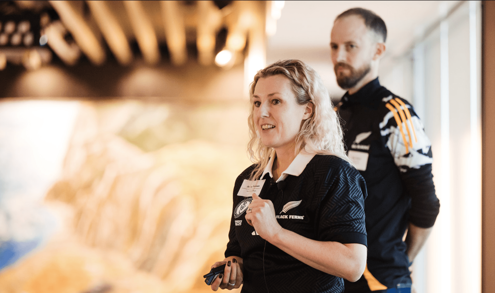 A blonde woman in a black Black Ferns polo shirt speaks, with a bearded man in an Adidas shirt standing behind her.