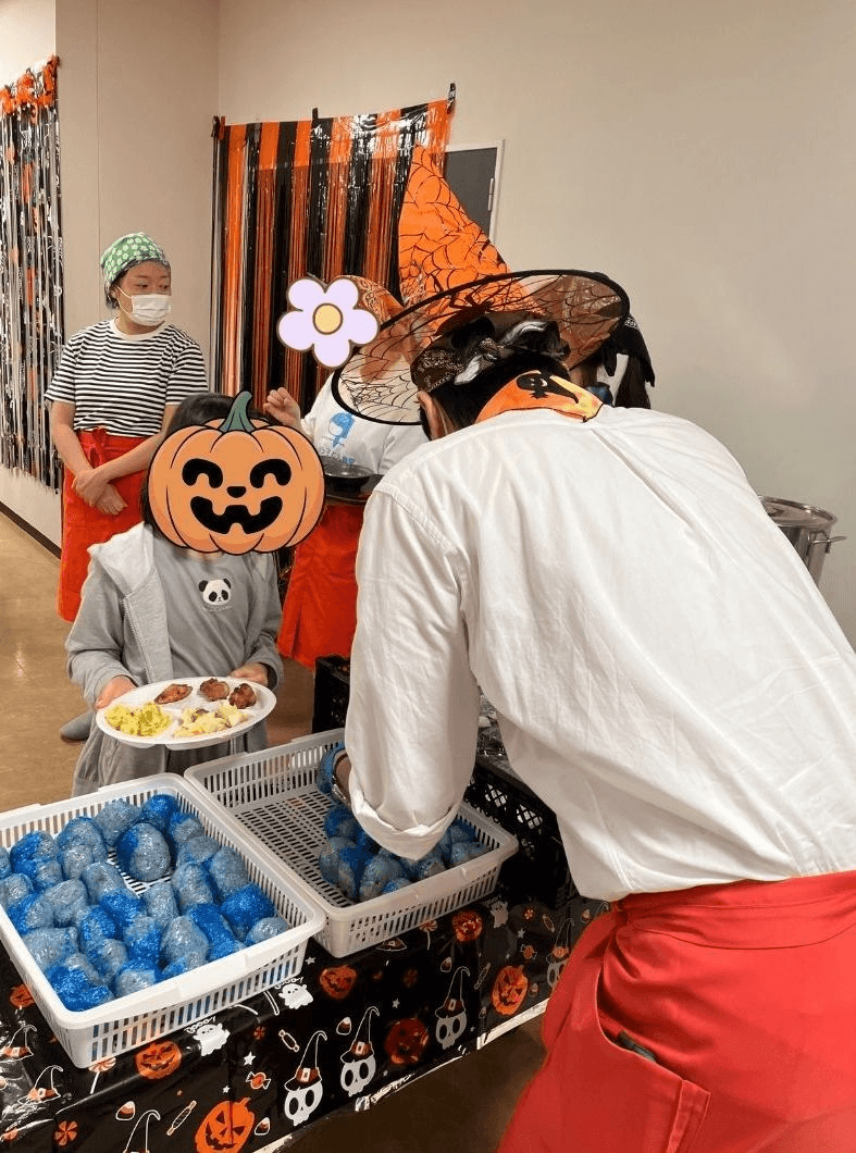 At a Halloween party, an adult in a witch hat serves food to a child with a pumpkin emoji over their face, next to baskets of blue rice balls.