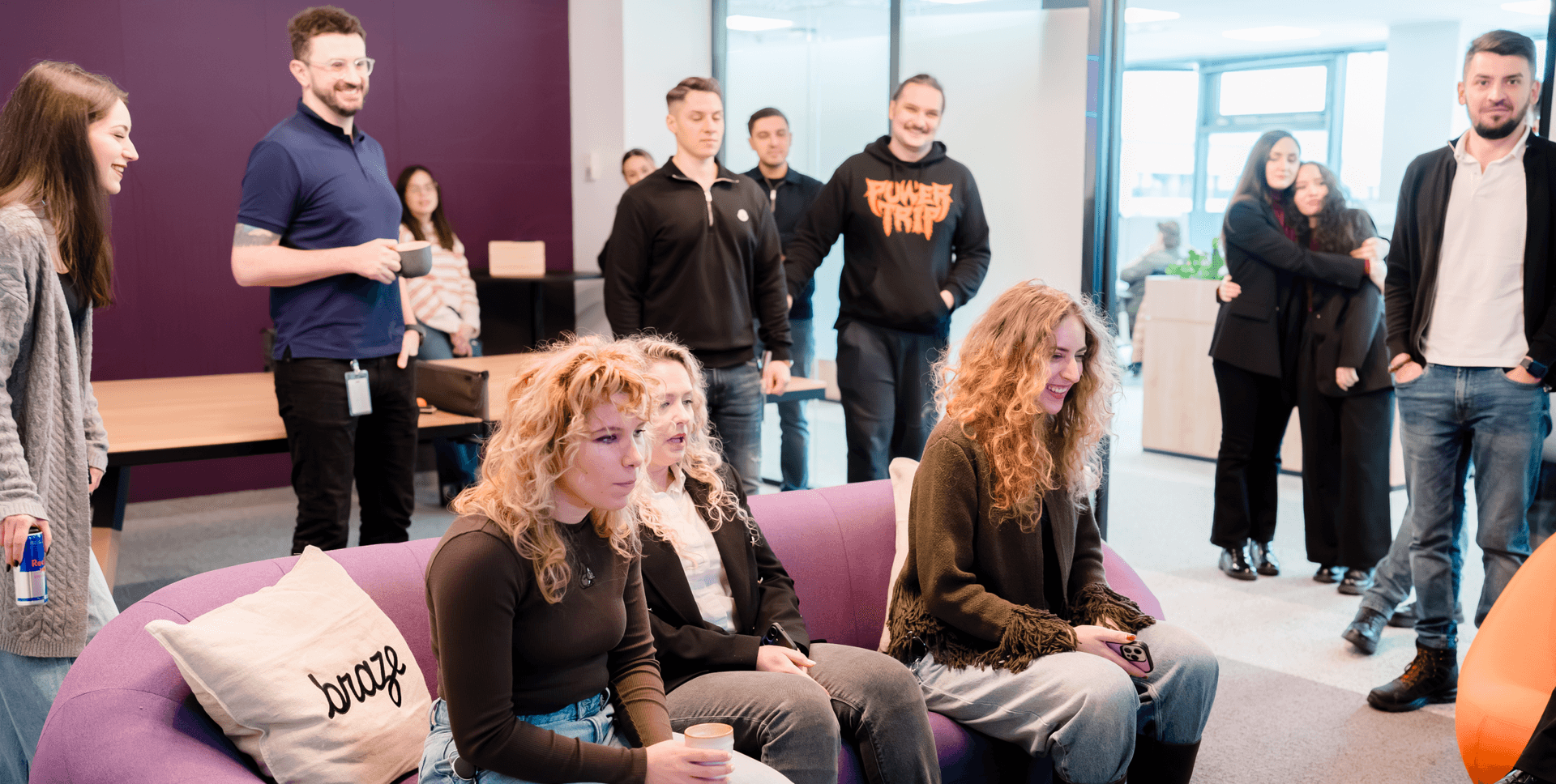 People gathered in an office lounge, some on a purple couch with a "braze" pillow, others standing.