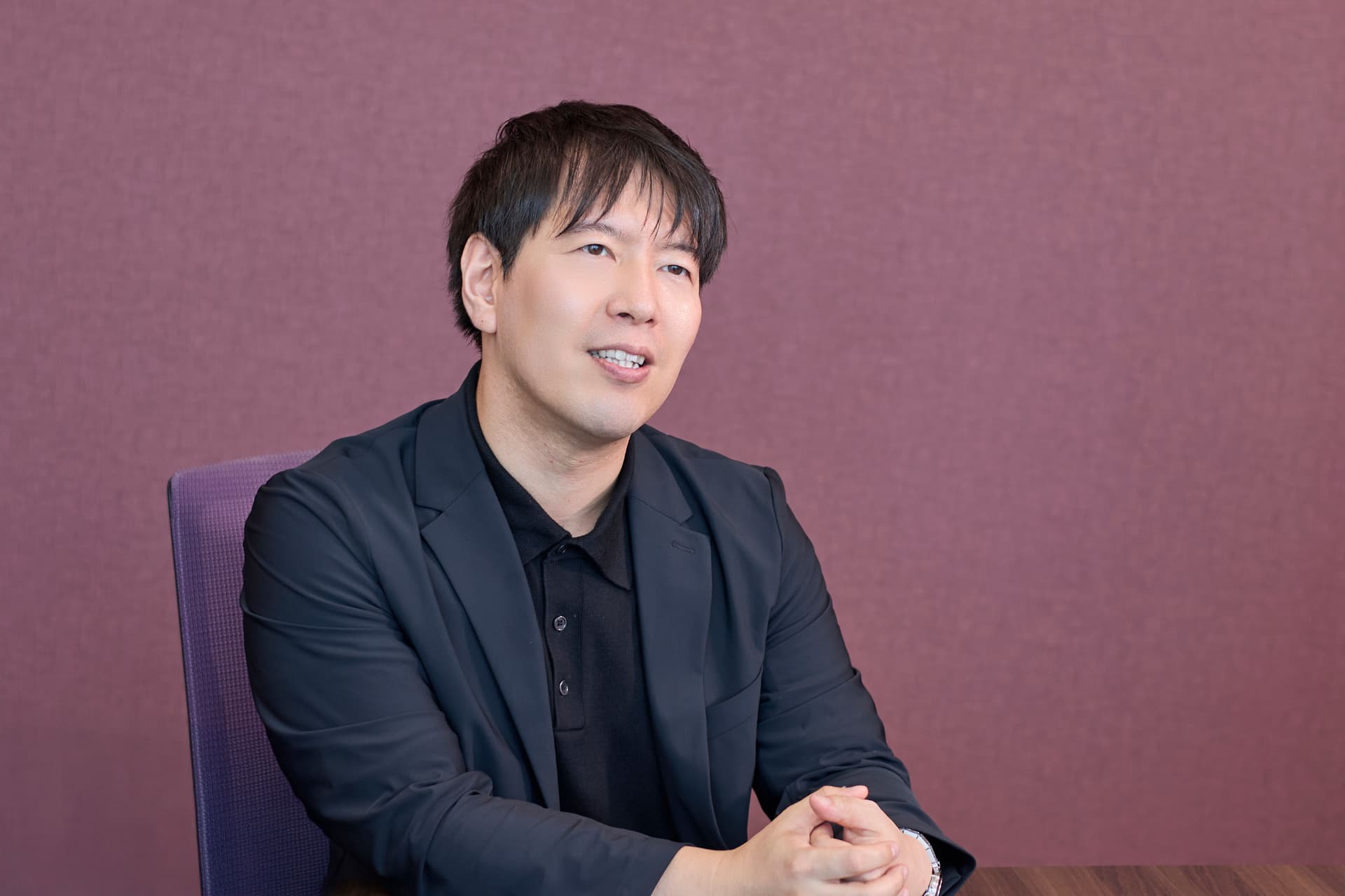 a man in a suit and black shirt is sitting at a table with his hands folded .