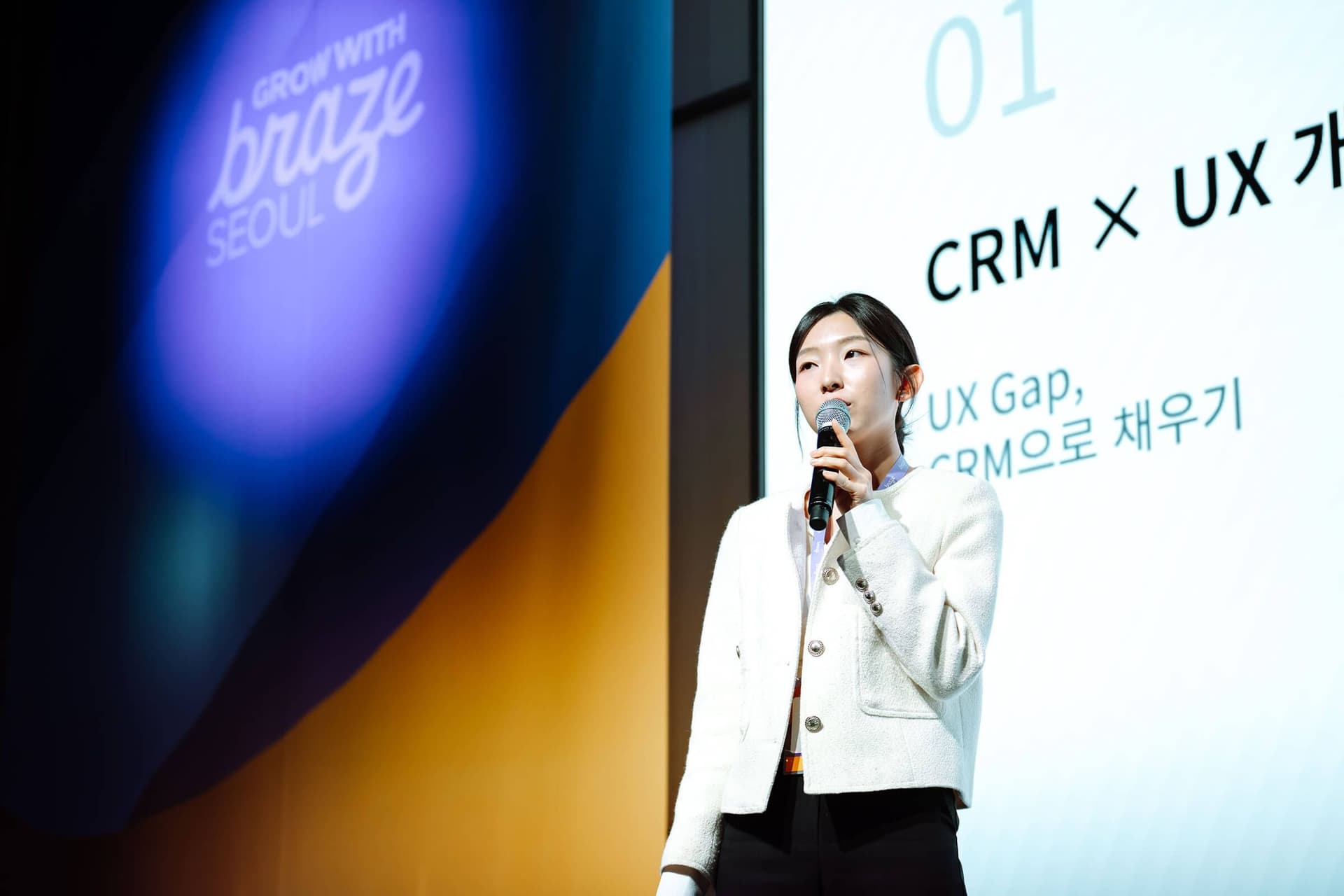 A woman presents on stage at a Braze Seoul event, with a screen showing "CRM x UX" text behind her.