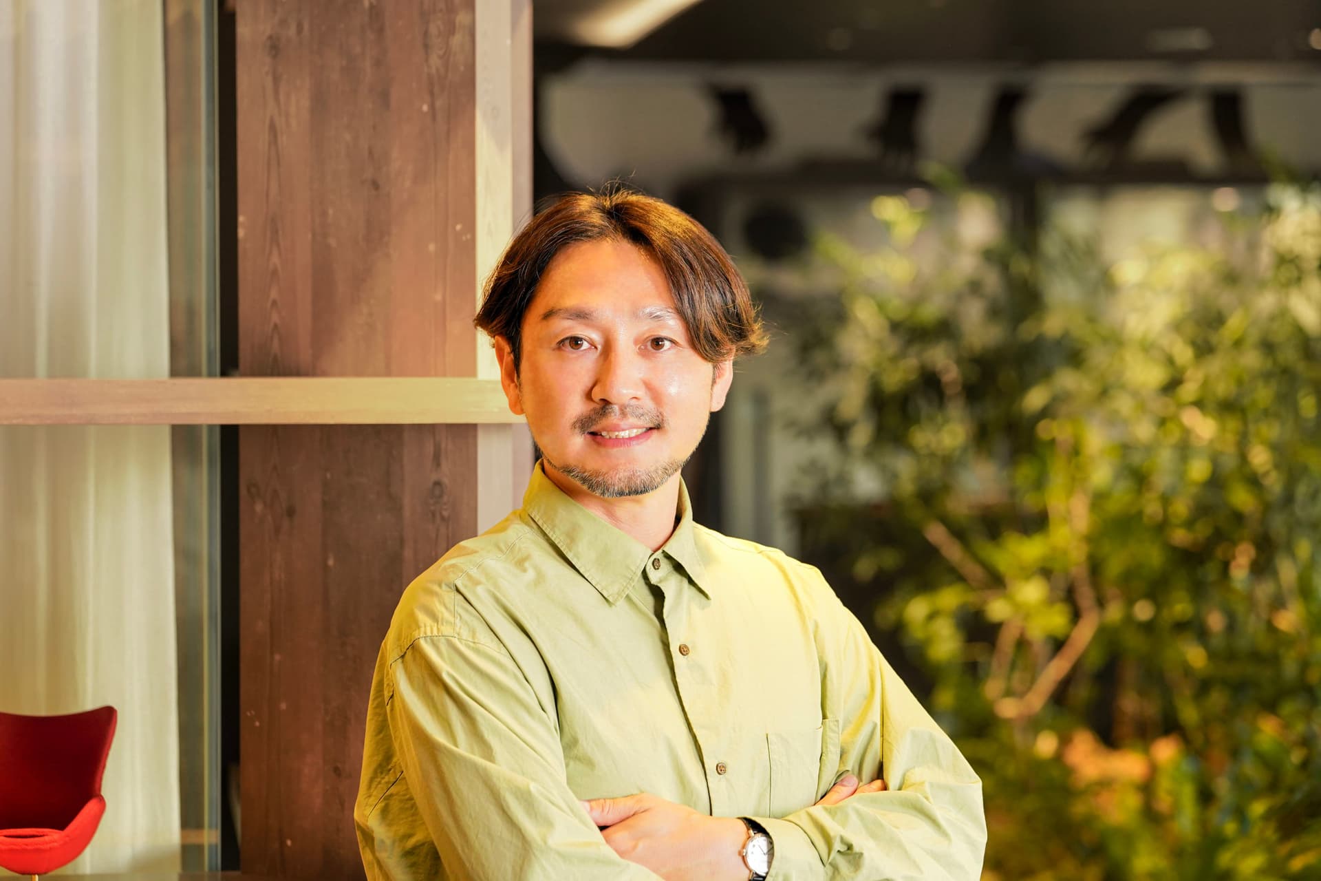 A smiling man with a beard, wearing a light green shirt, stands with his arms crossed indoors.