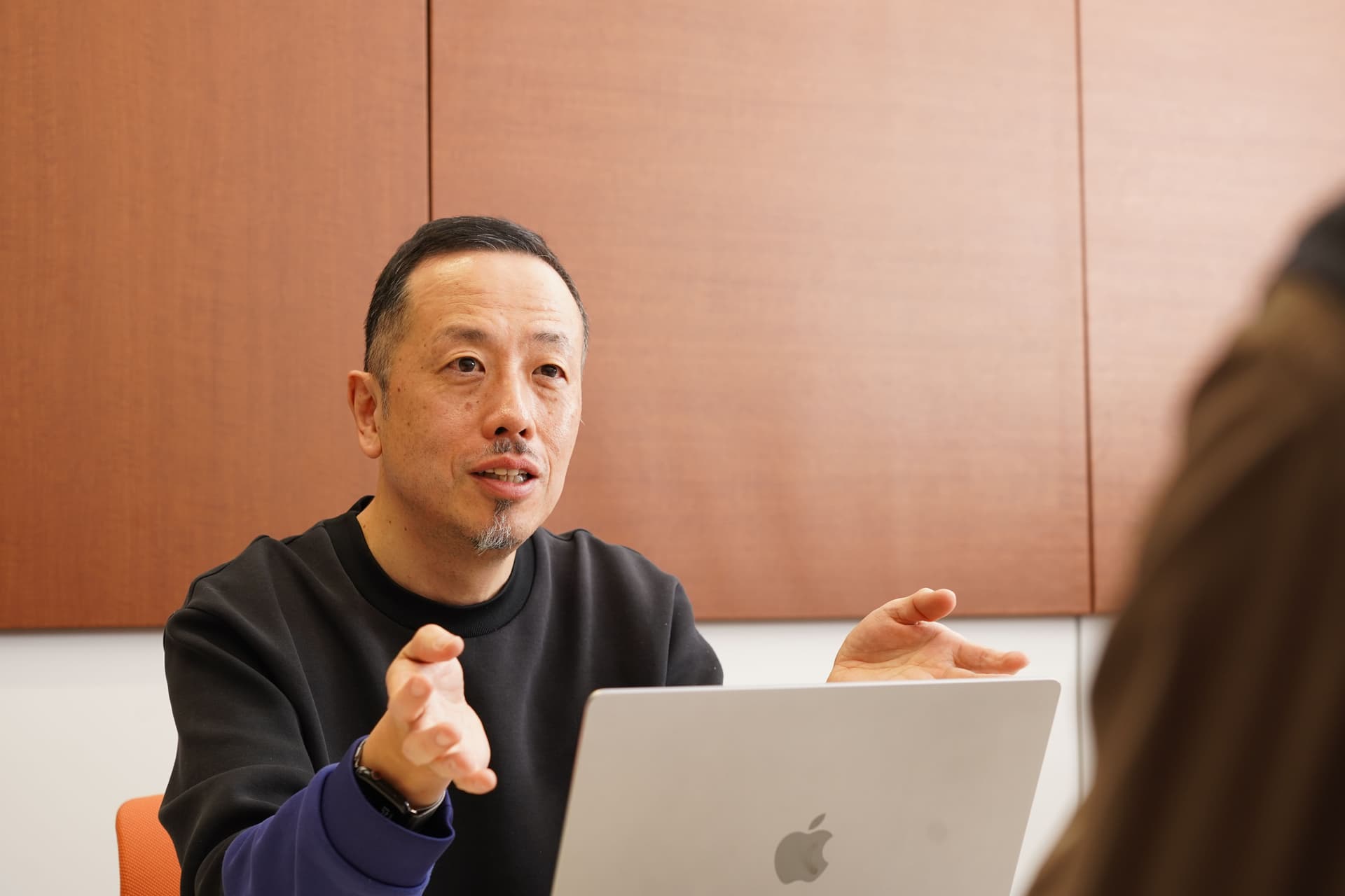 あごひげを生やした東アジア系の男性が話しながら身振り手振りをし、目の前のテーブルにはノートパソコンが置かれている。