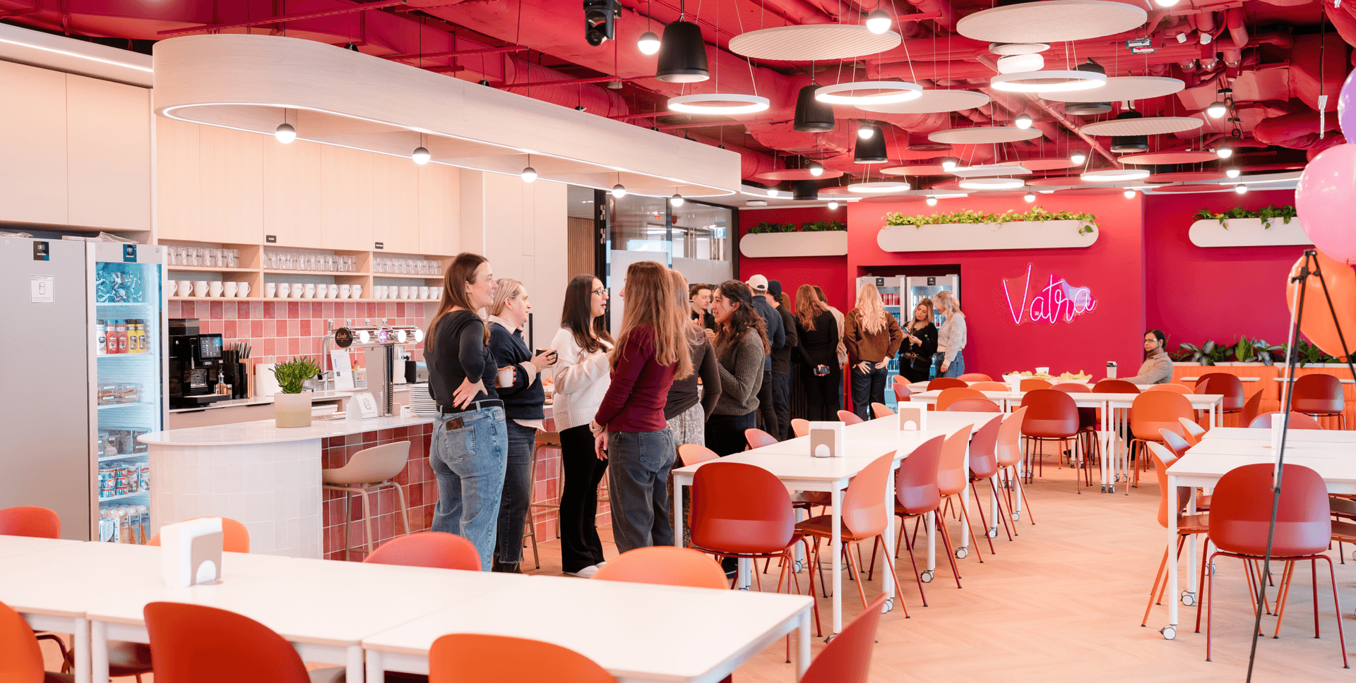 A modern, vibrant office cafeteria with pink walls, orange chairs, a kitchenette, and people socializing.