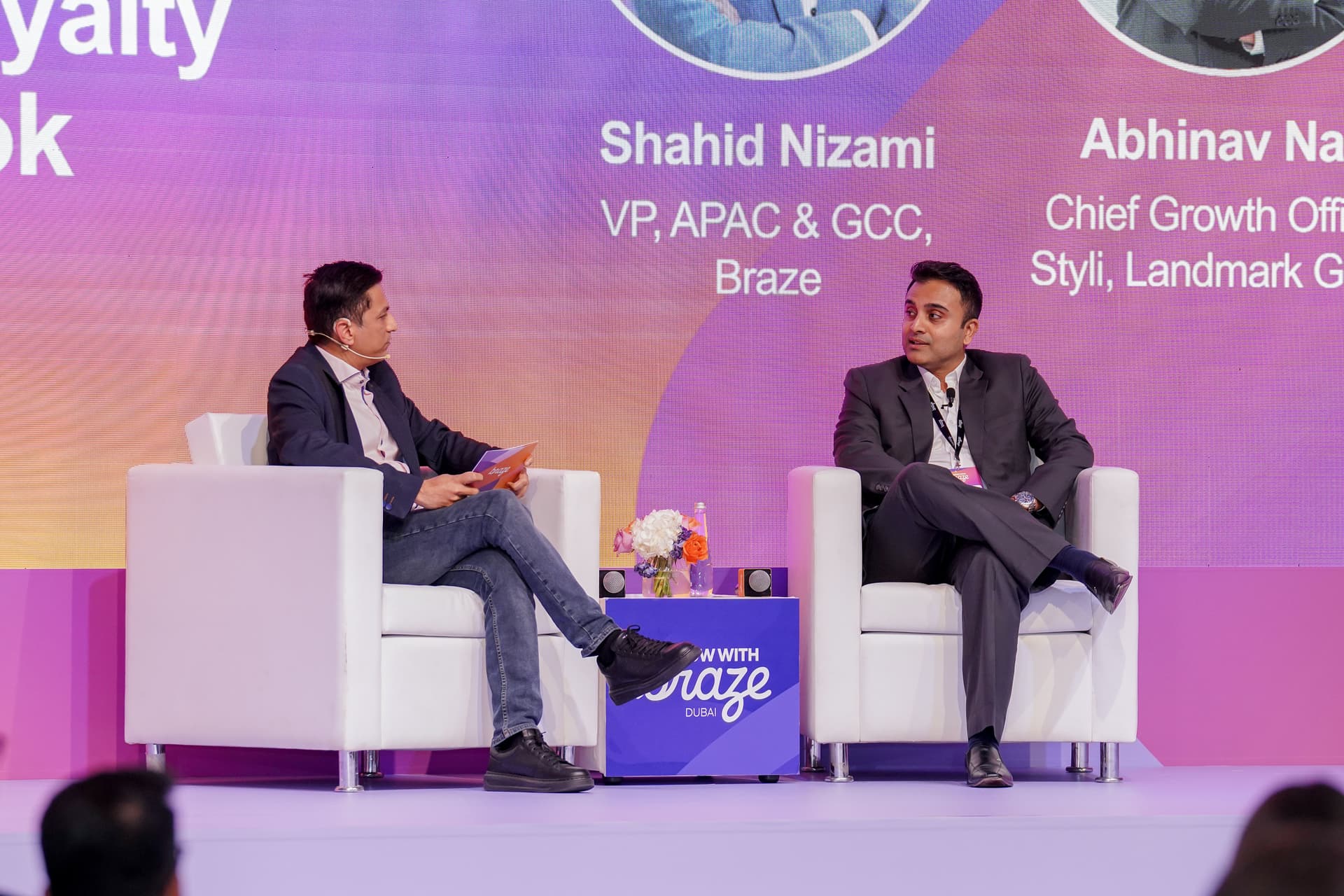 Two men seated on a stage, participating in a panel discussion with names and titles on the backdrop.