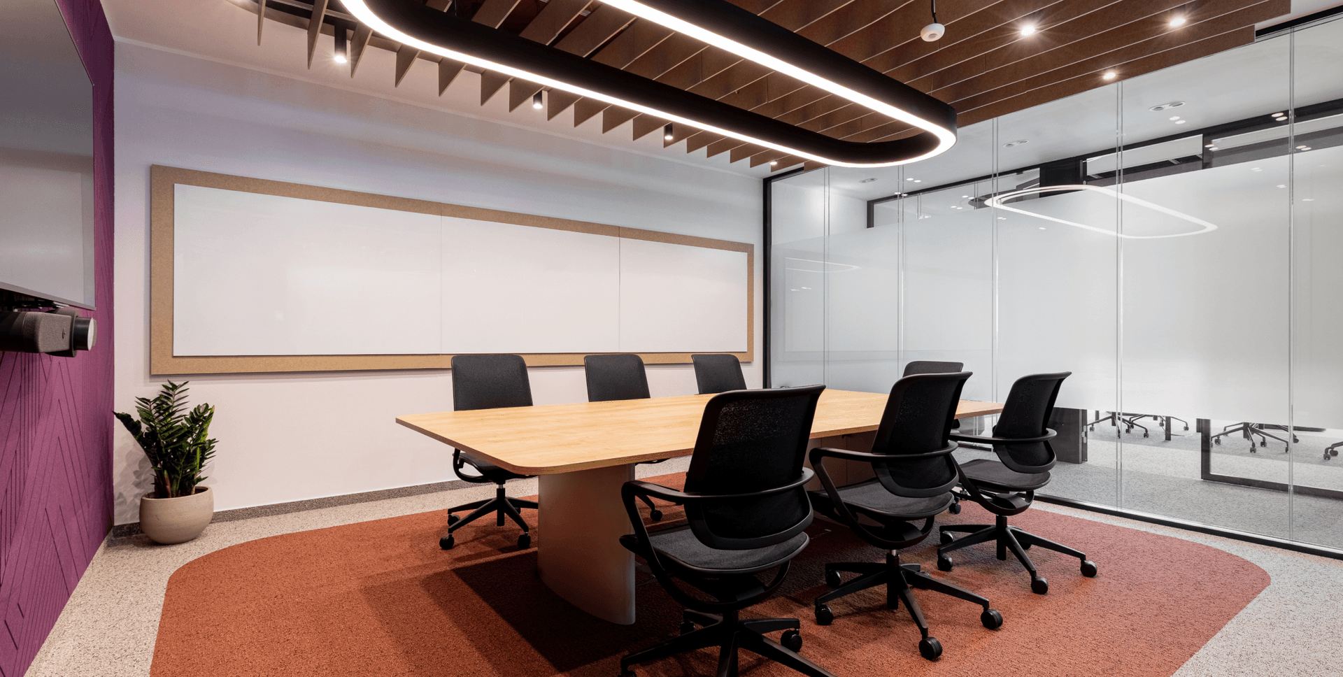 A modern meeting room with a light wood conference table, black chairs, large whiteboard, and purple and terracotta accents.